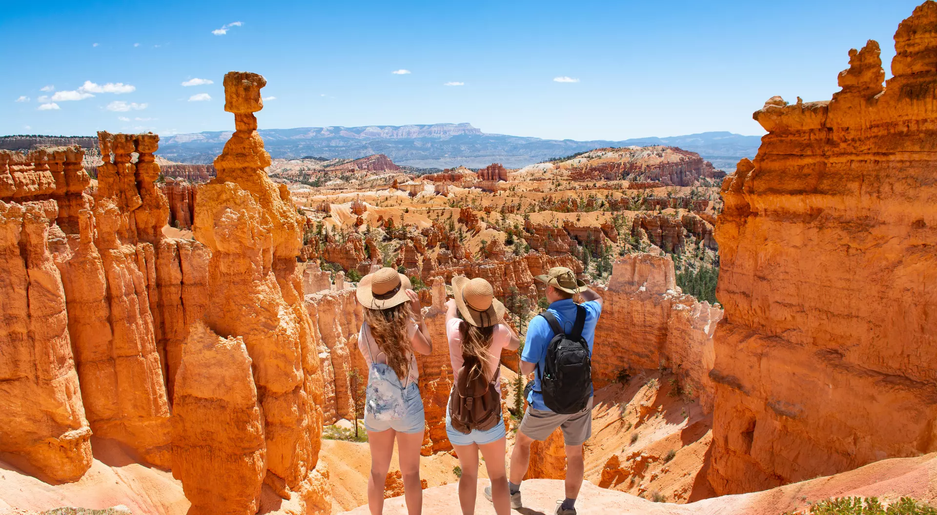 Three travelers stand at the edge of a canyon, gazing at orange rock formations under a clear blue sky.