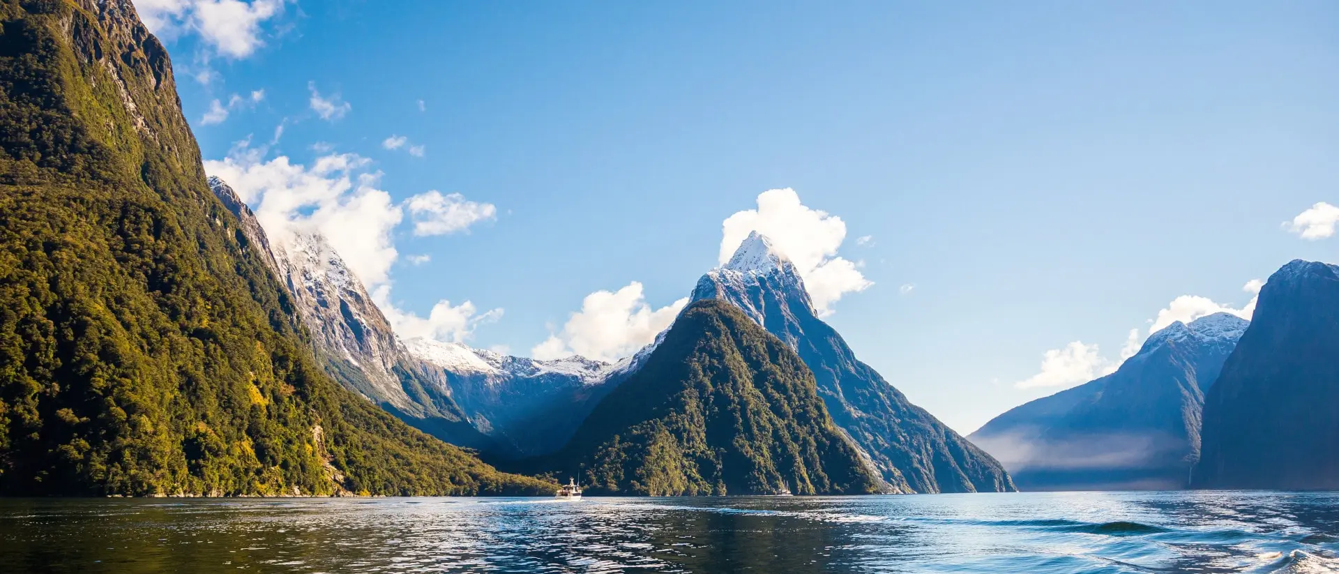 Milford Sound, New Zealand Towering forested peaks, snowy mountains, and calm blue waters under a clear sky in New Zealand’s dramatic Milford Sound fjord.