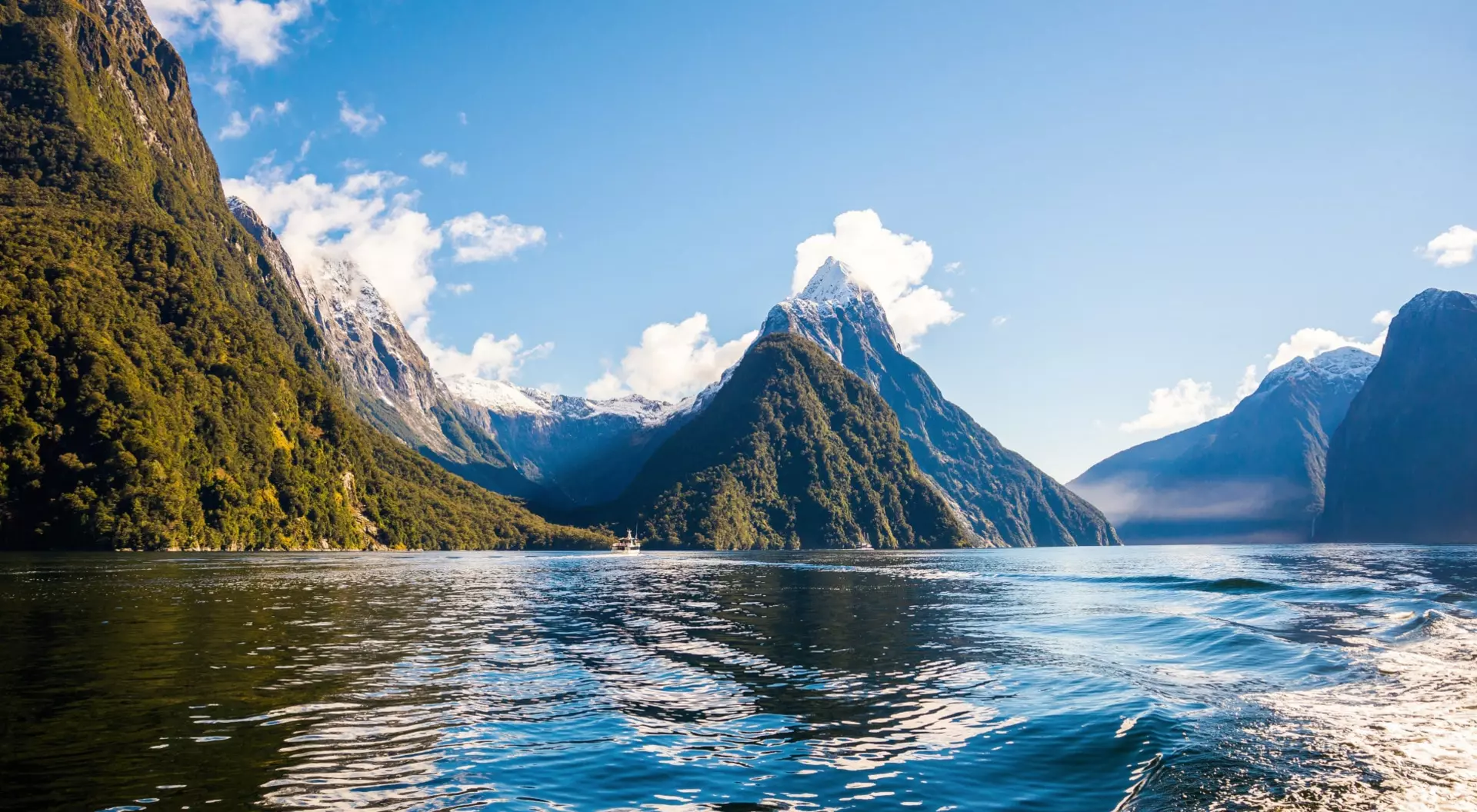 Towering forested peaks, snowy mountains, and calm blue waters under a clear sky in New Zealand’s dramatic Milford Sound fjord.