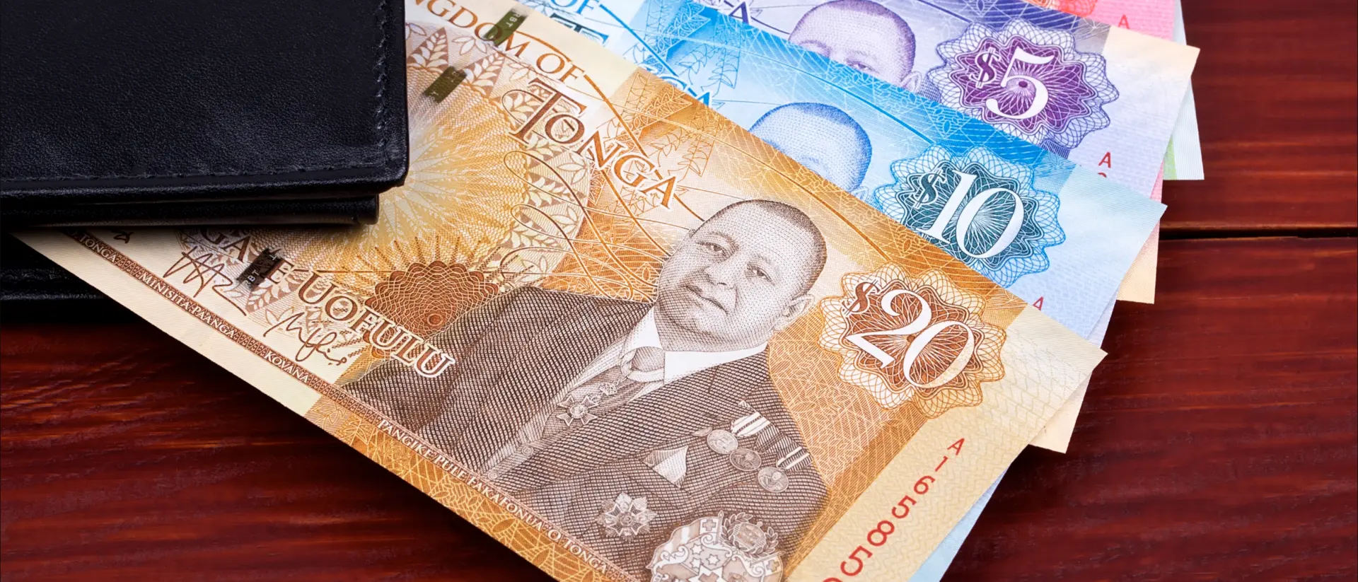 A wallet with Tongan banknotes in various denominations displayed on a wooden surface.