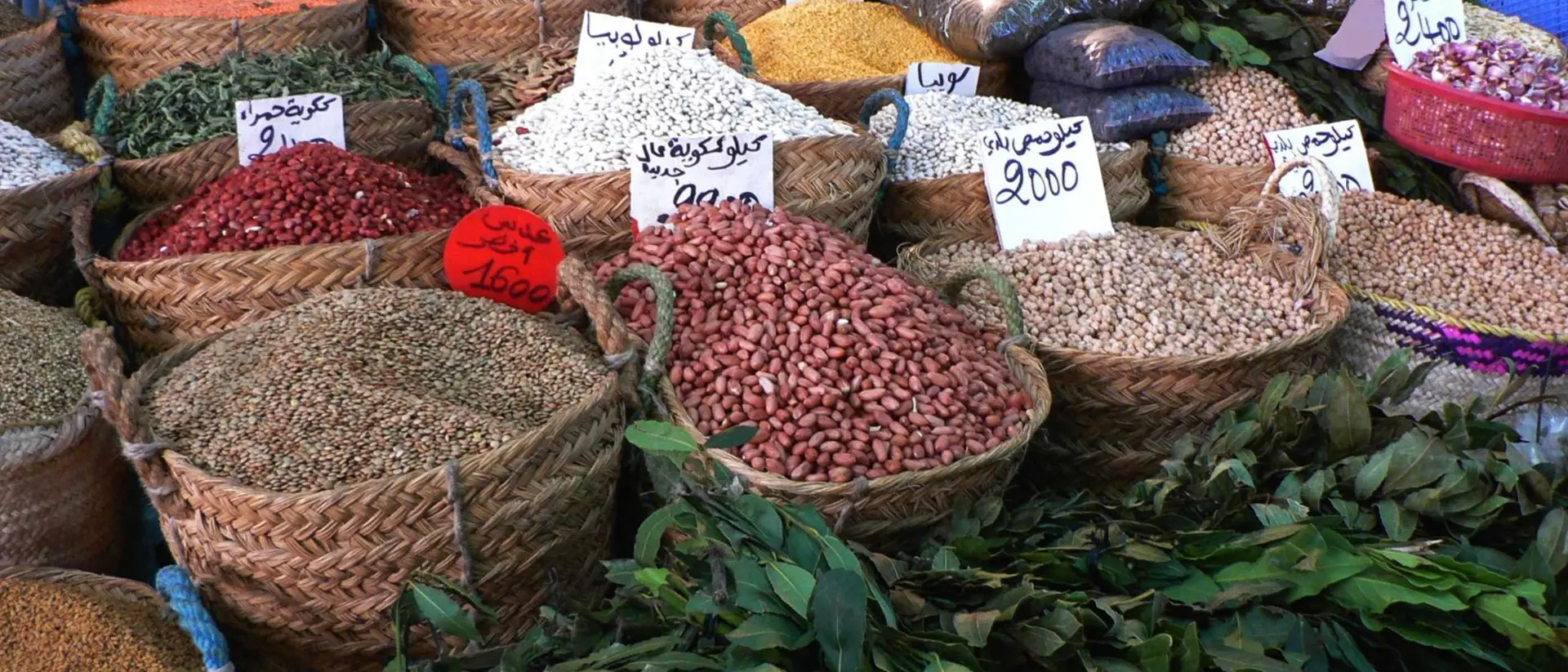 Baskets of colorful spices and legumes arranged in a market, with price tags visible in Arabic.