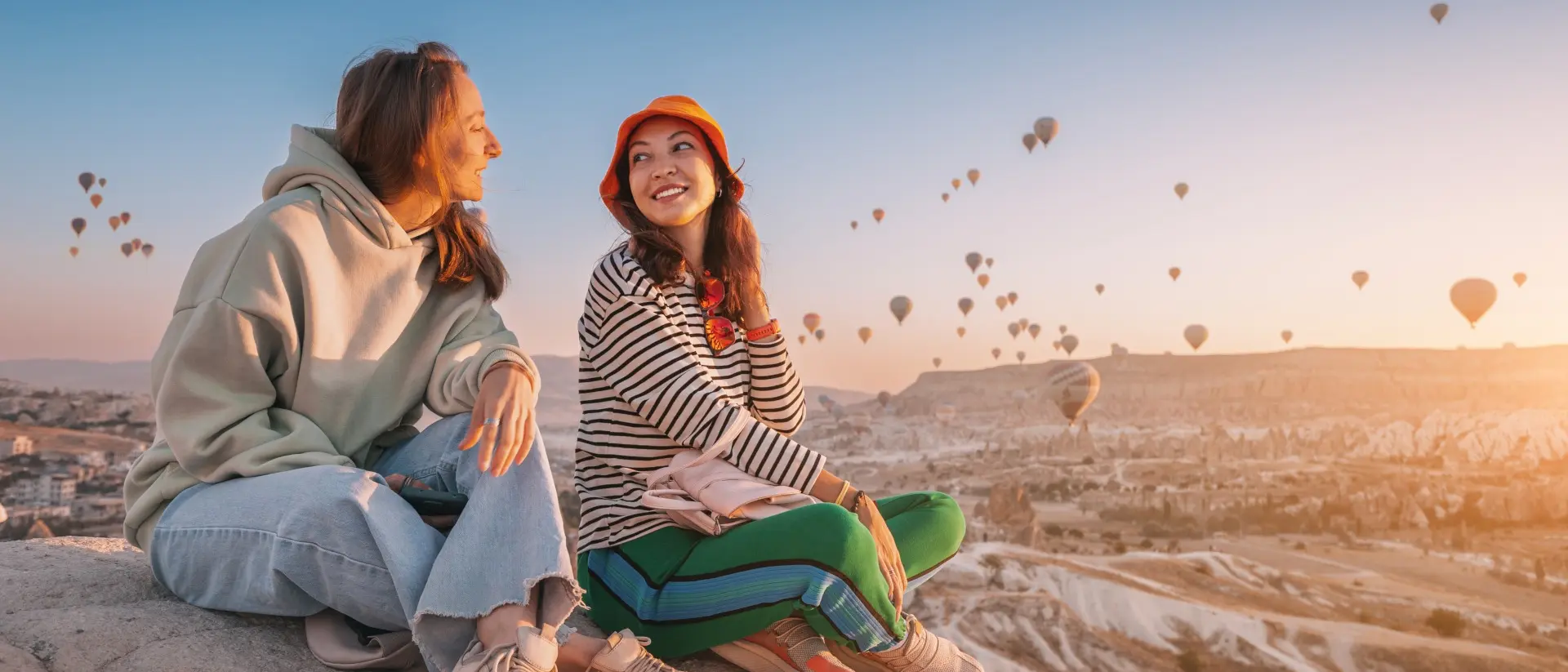 Two women sit on a cliff overlooking hot air balloons in Cappadocia, Turkiye.