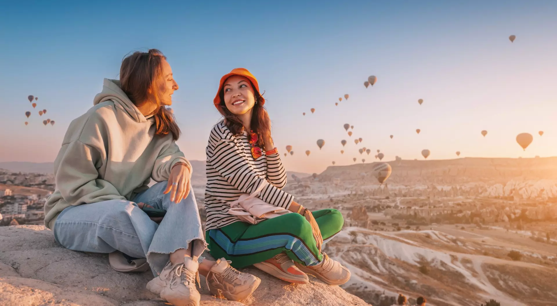 Two women sit on a cliff overlooking hot air balloons in Cappadocia, Turkiye.