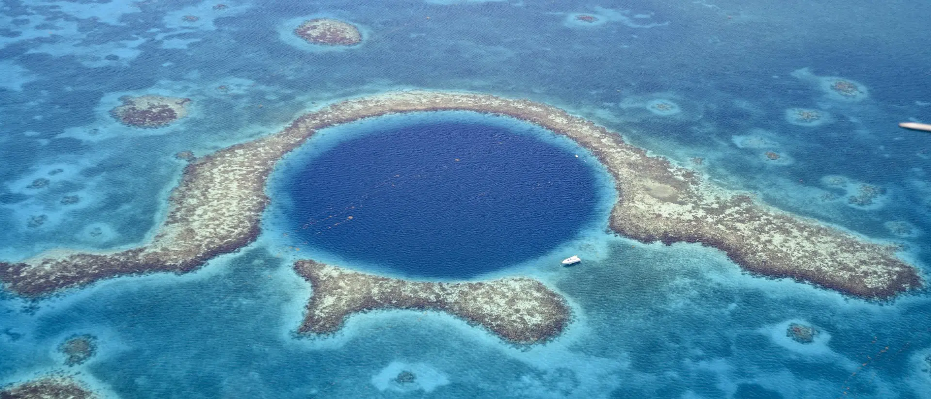 Aerial view of Great Blue Hole a marine sinkhole and geological wonder in the Belize Barrier Reef Reserve. A small white yacht is moored at it's edge