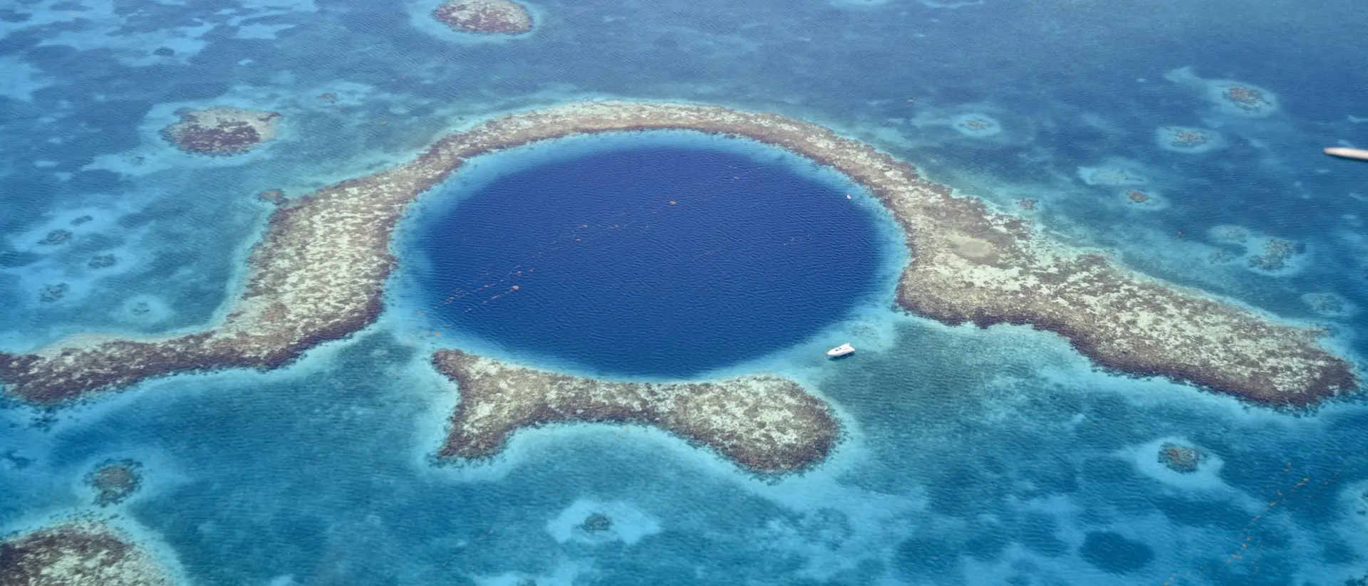Aerial view of Great Blue Hole a marine sinkhole and geological wonder in the Belize Barrier Reef Reserve. A small white yacht is moored at it's edge