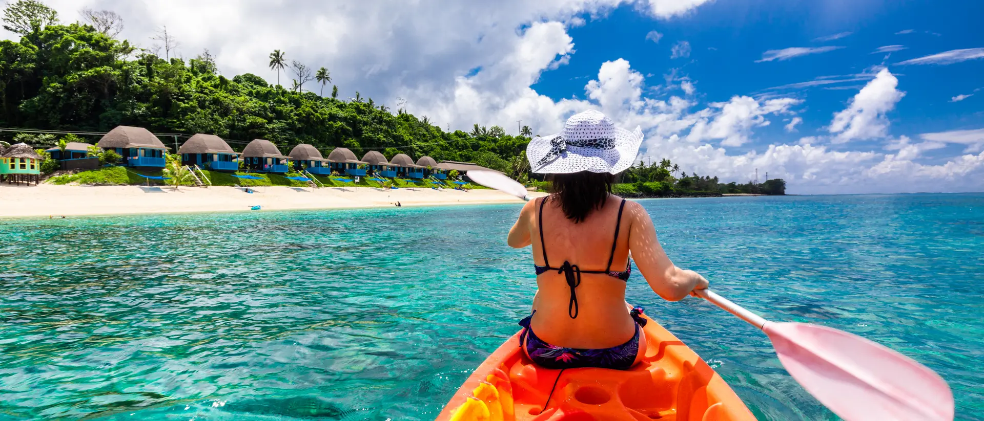 woman kayaking in waters off a beach with bungalows