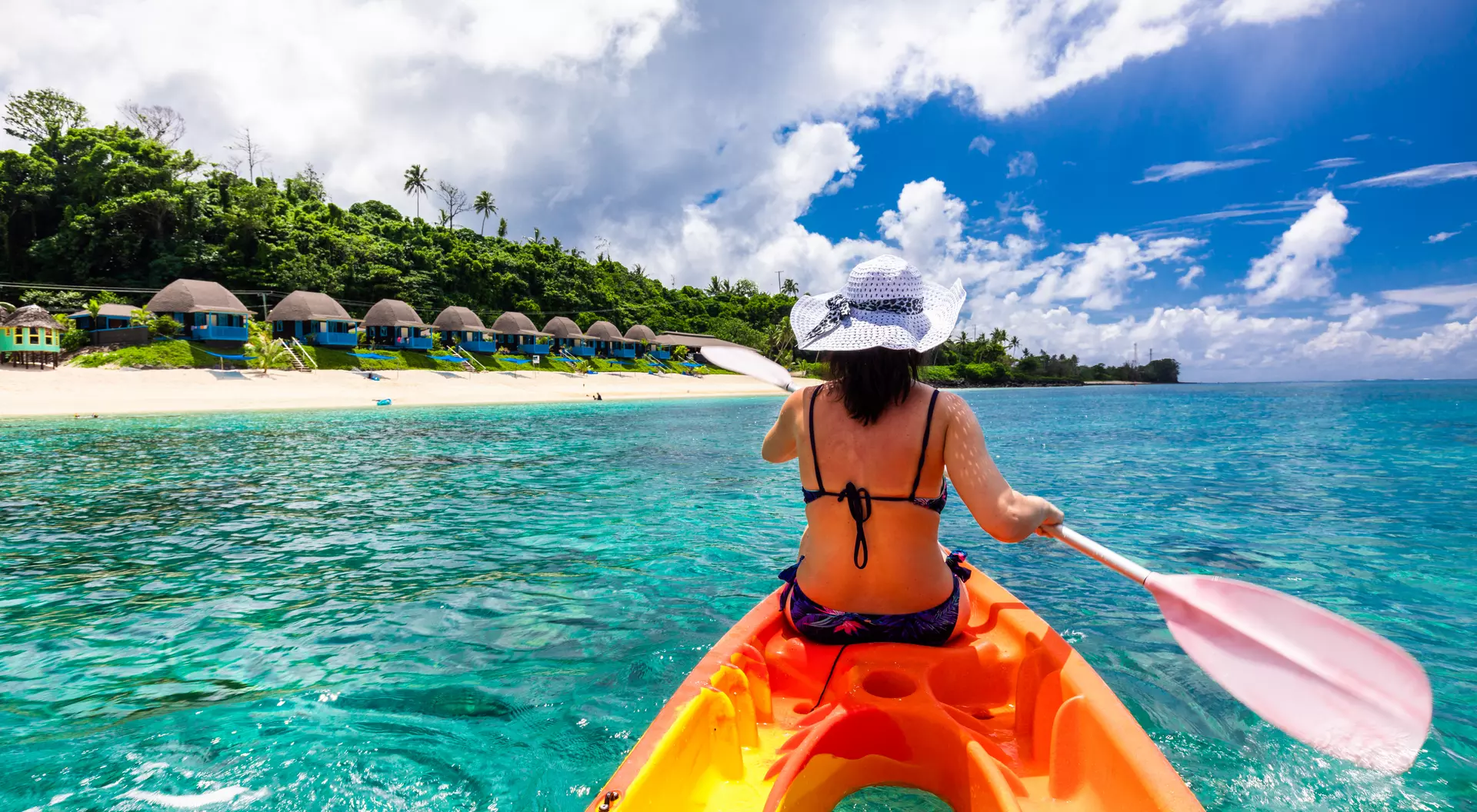 woman kayaking in waters off a beach with bungalows