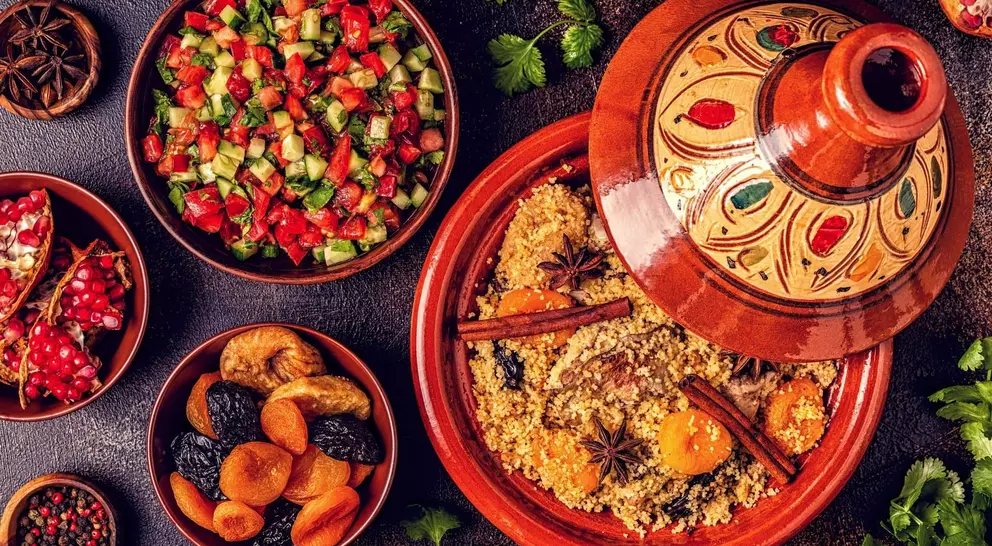 A colorful spread of couscous with spices, surrounded by salads, dried fruits, and garnishes on a textured surface.