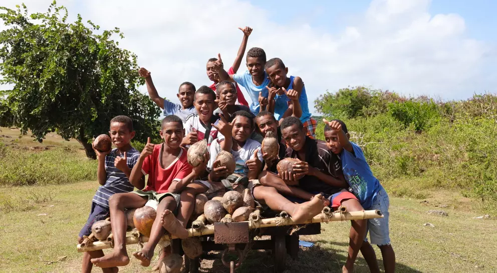 A group of joyful boys poses on a wooden cart, holding coconuts outdoors in a lush green setting.