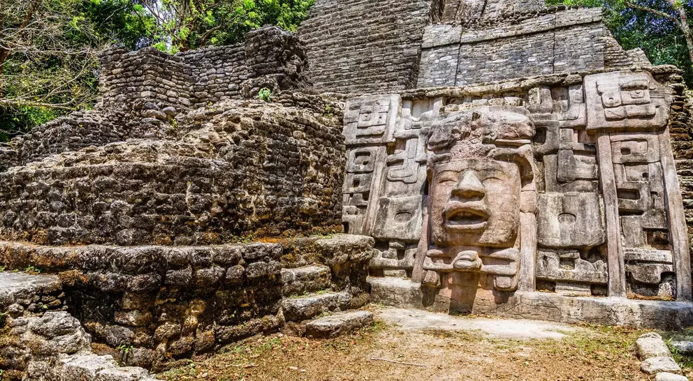 Ancient stone ruins with a carved face, surrounded by greenery and blue sky.