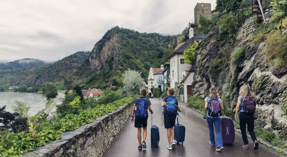 Travelers with suitcases walk along a scenic road surrounded by mountains and greenery near a riverbank.