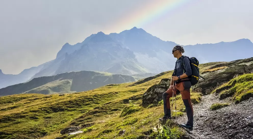 Hiker standing on a rocky trail, gazing at mountains and a rainbow in the sky.