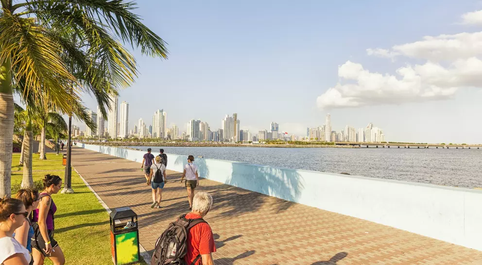 People walking along a waterfront promenade with a skyline and palm trees in the background under a clear sky.