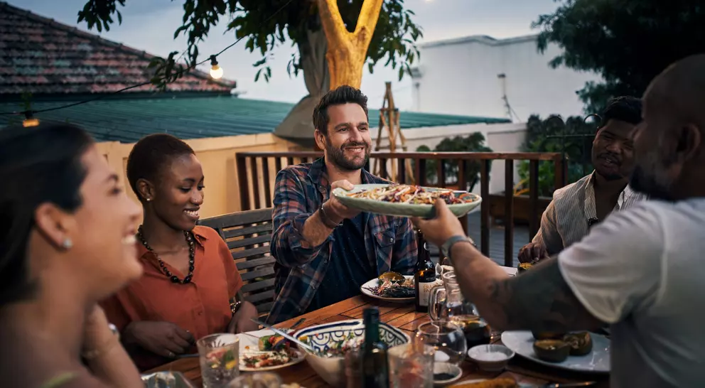 A group of friends enjoys dinner outdoors, sharing food and smiling at each other under string lights.