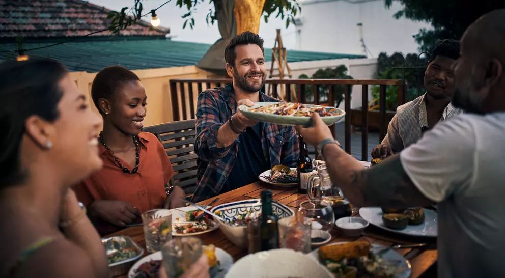 A group of friends enjoys dinner outdoors, sharing food and smiling at each other under string lights.