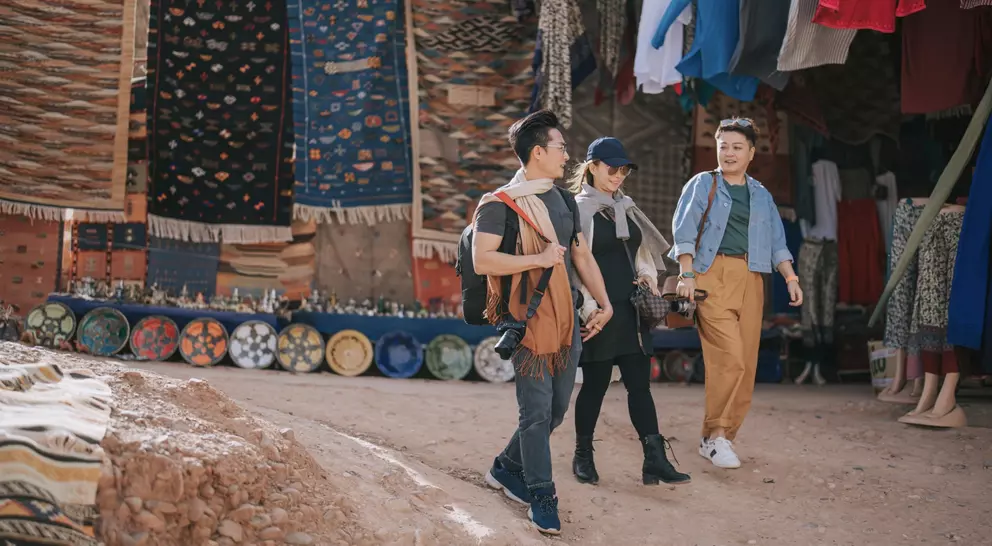 Three people walk through a vibrant market, surrounded by colorful textiles and handmade pottery.
