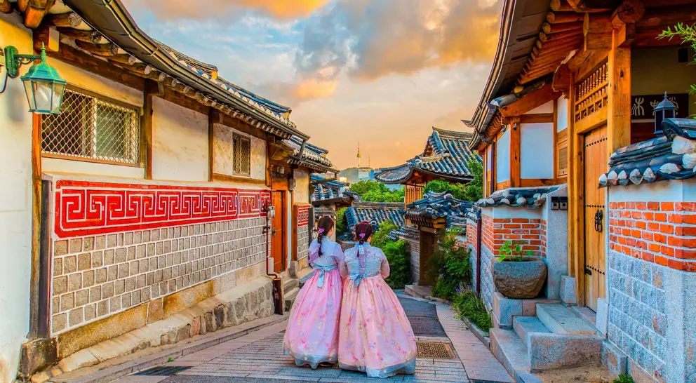 Two girls in traditional attire stroll through a picturesque alley lined with wooden houses and colorful sky at sunset.