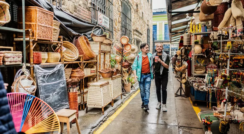 Two men stroll through a vibrant market alley filled with colorful handmade crafts and woven baskets.