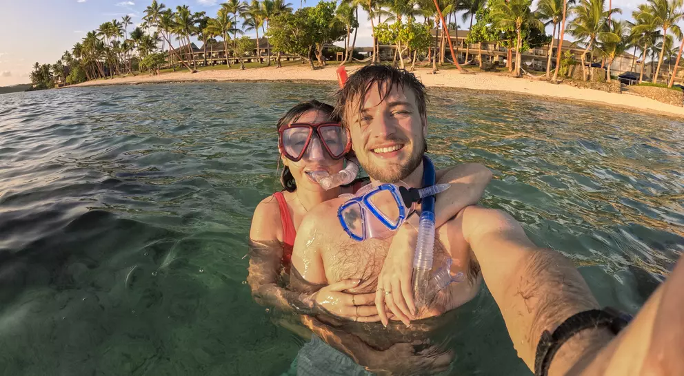 A waist up selfie of a young couple snorkeling in the sea at sunset on a tropical resort in Fiji. There are palm trees along the beach behind them. The couple are smiling into the camera and showing happy emotions.