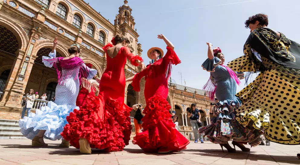 A group of women in colorful flamenco dresses dance in a plaza with historic architecture in the background.