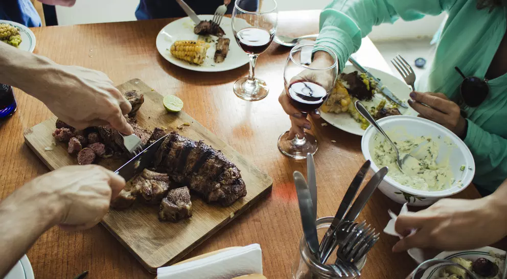 A wood table laden with plates of food, glasses of wine, and hands sharing a meal together.
