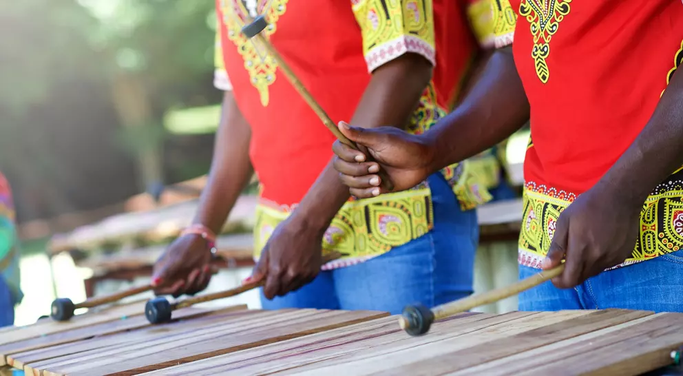 Three musicians in colorful shirts play a wooden xylophone outdoors, holding mallets and striking the bars.