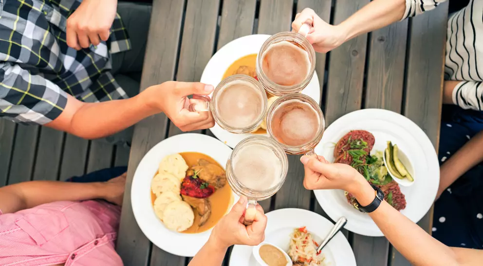 Friends toasting with beer mugs over a table of diverse dishes, enjoying a meal together.