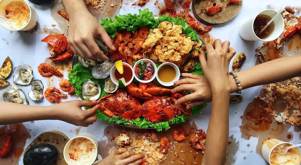 A feast of seafood, including lobsters and oysters, with hands reaching for food on a messy table covered in shells and sauces.