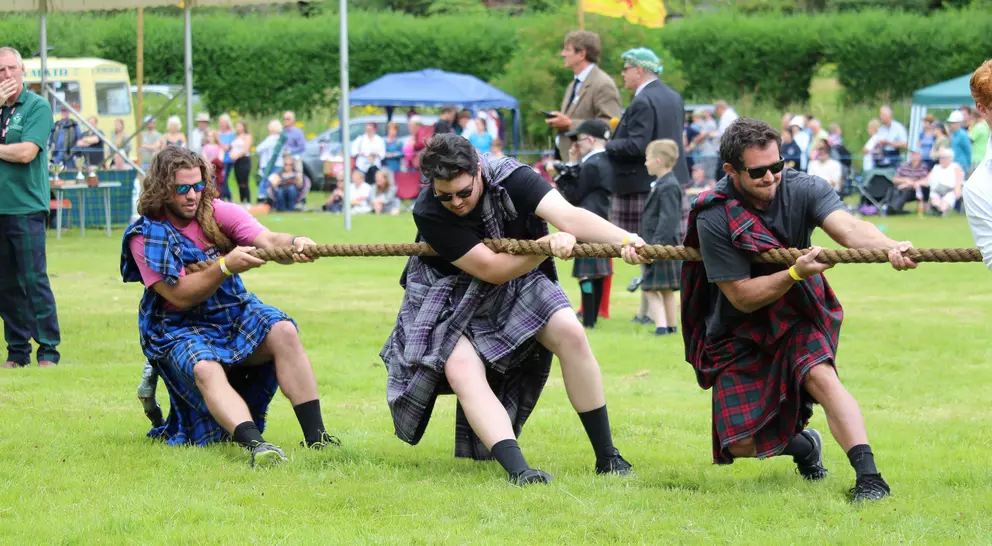 Three men in kilts are pulling a rope in a tug-of-war competition, with a crowd watching in the background.