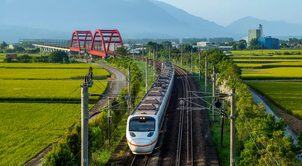 A train travels through a green landscape with rice fields and a red bridge in the background, under a clear blue sky.