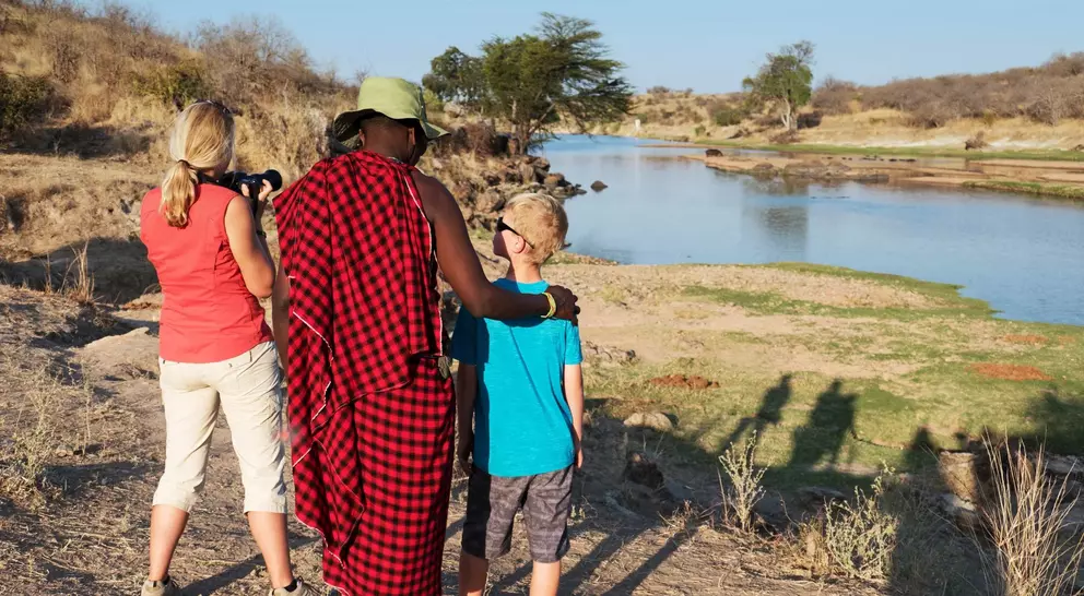 A woman and a boy watch a river with a man in traditional attire, capturing the moment as the sun sets.
