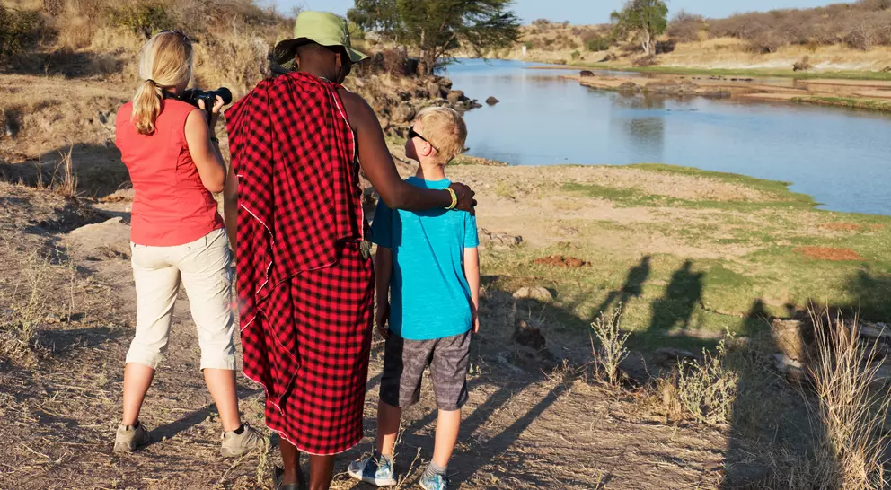 A woman and a boy watch a river with a man in traditional attire, capturing the moment as the sun sets.