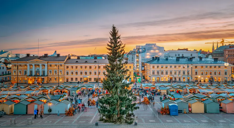 A vibrant market square at sunset, featuring a large Christmas tree and festive stalls filled with visitors and decorations.