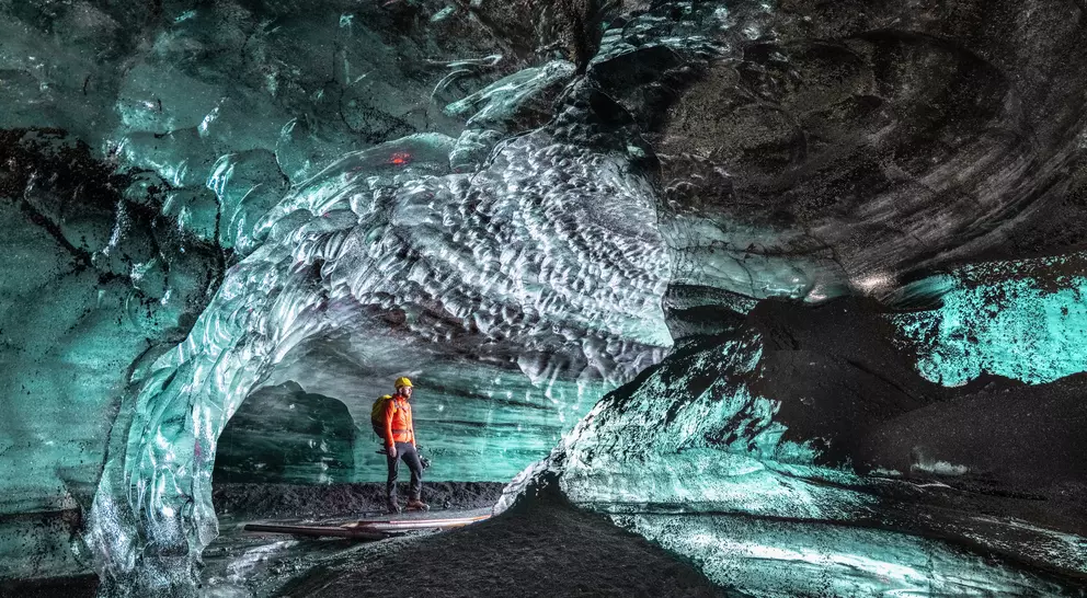A person in an orange jacket stands inside a stunning ice cave with textured blue ice formations and intricate patterns.