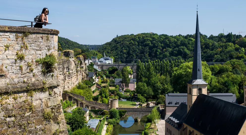 A person sits on a fortress wall overlooking lush greenery and a river, with buildings and a church spire visible below.