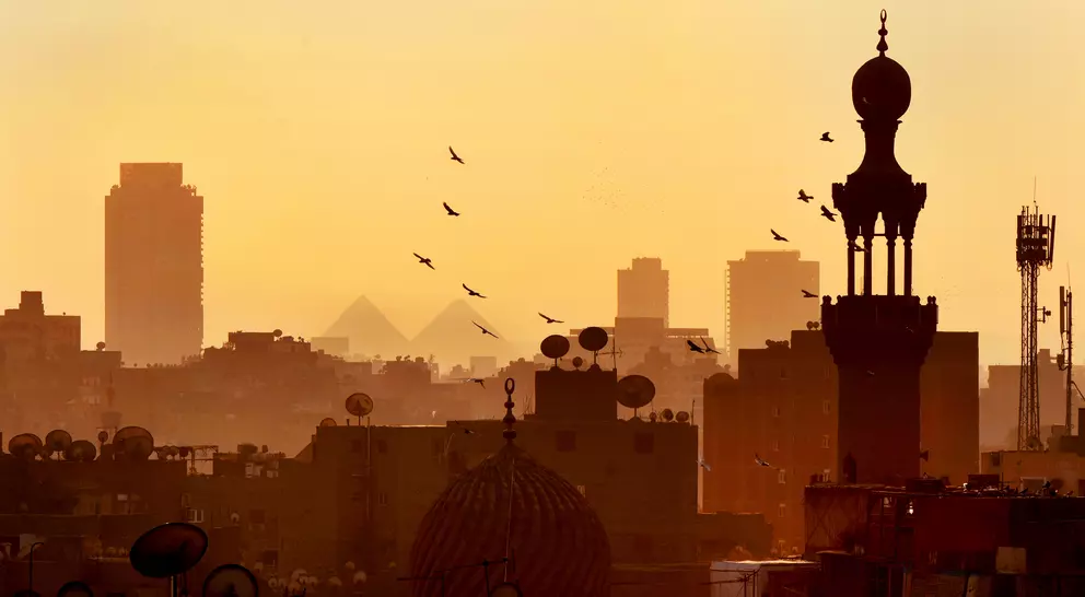 The skyline of Cairo at sunset with birds over domes and satellite dishes with distant pyramids at Giza