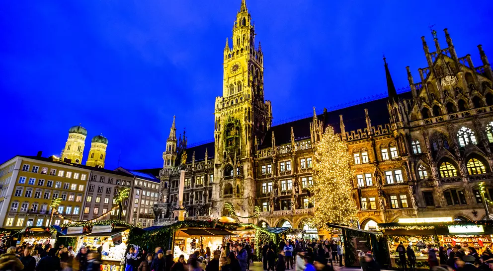 A bustling market scene at night with a tall clock tower and a lit Christmas tree in the background.