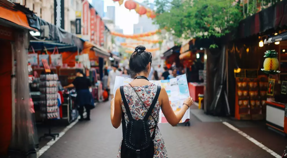 A woman stands in a market holding a map, surrounded by colorful stalls and hanging lanterns.