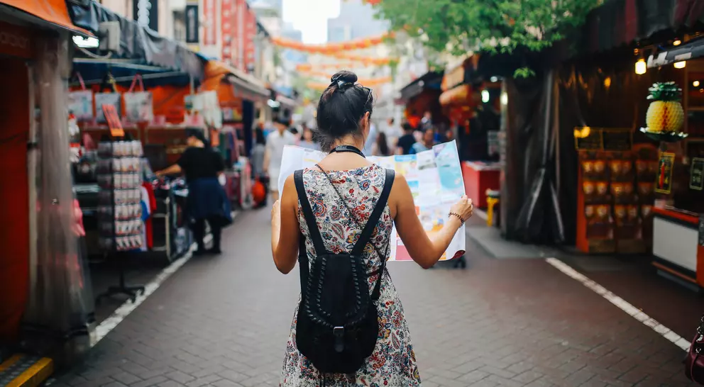 A woman stands in a market holding a map, surrounded by colorful stalls and hanging lanterns.
