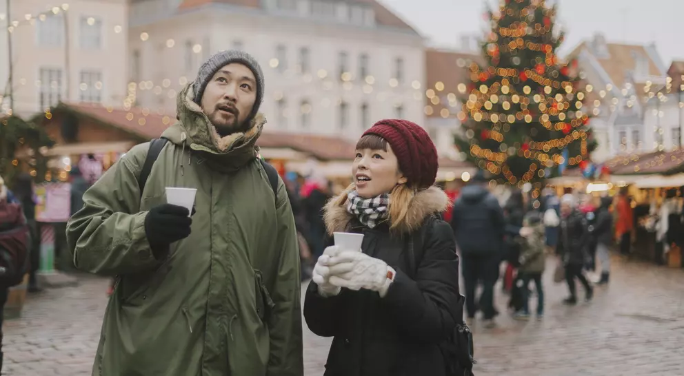 A man and woman look up, holding cups, in a festive market with holiday lights and a Christmas tree in the background.