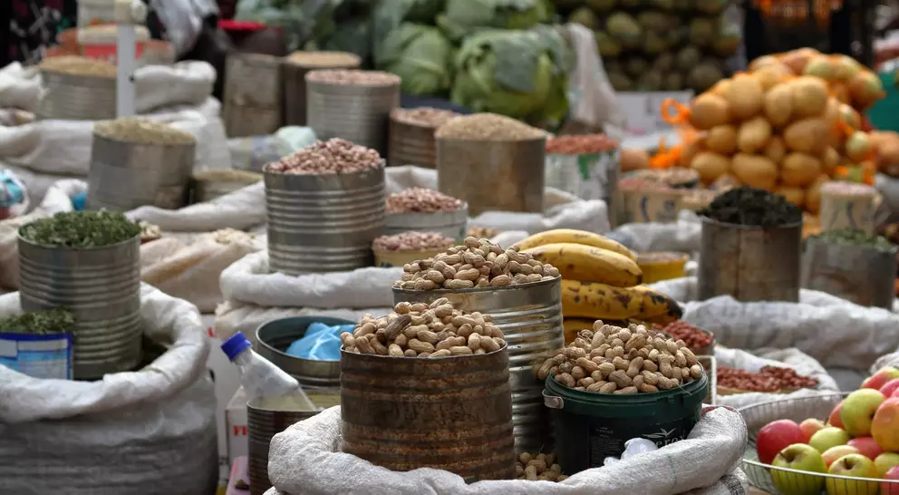 Various sacks of nuts and legumes are arranged in a bustling market, with fruits and vegetables in the background.