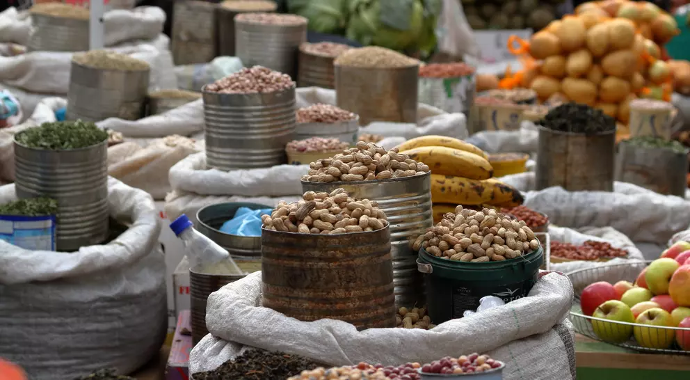 Various sacks of nuts and legumes are arranged in a bustling market, with fruits and vegetables in the background.