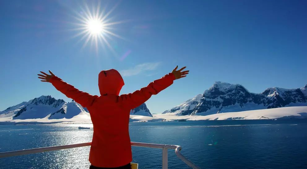 Person in a red jacket spreading arms wide, facing sunlit mountains and ocean under a clear blue sky.