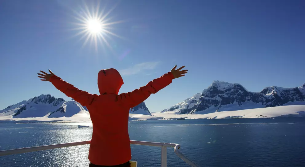 Person in a red jacket spreading arms wide, facing sunlit mountains and ocean under a clear blue sky.