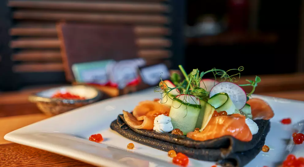 A gourmet dish with black crepes, smoked salmon, cucumber, cream, and orange caviar, elegantly plated on a white dish.