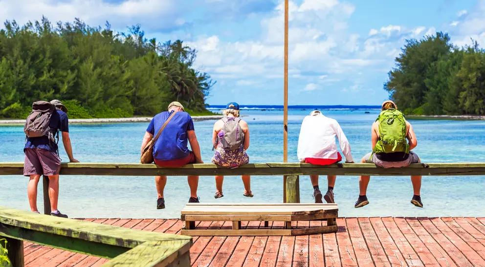 Five people sit on a wooden railing by the water, facing the ocean under a blue sky with scattered clouds.