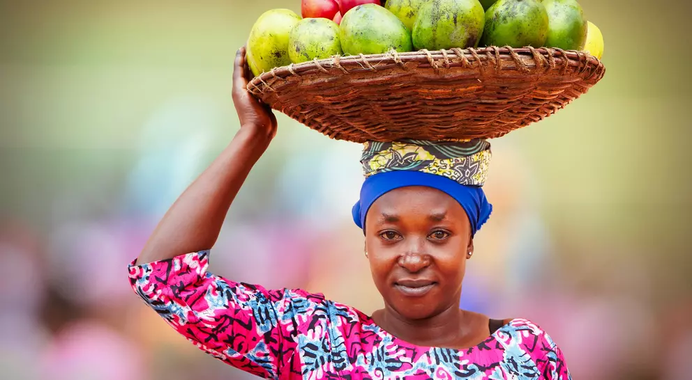 A woman in a colorful outfit balances a basket of fruits on her head while smiling at the camera.
