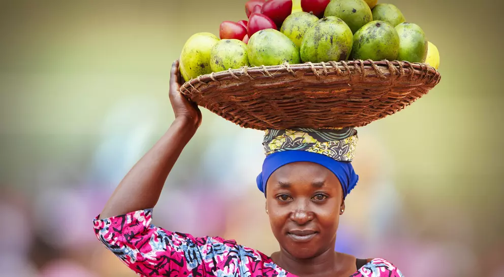 A woman in a colorful outfit balances a basket of fruits on her head while smiling at the camera.