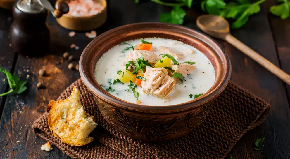 Bowl of creamy salmon soup with vegetables, garnished with herbs, beside a piece of bread on a woven mat.