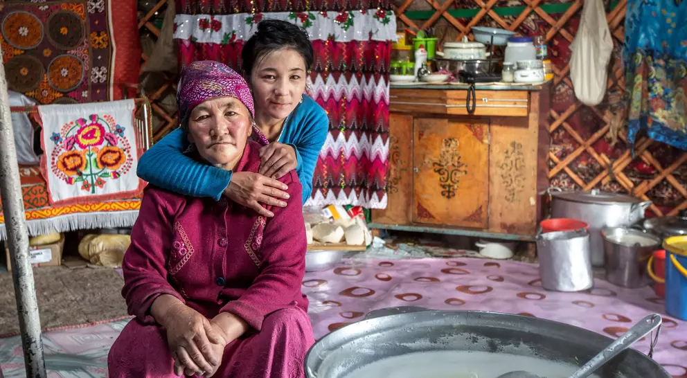 A young girl embraces an elderly woman in a cozy, traditional yurt adorned with colorful textiles and kitchen items.